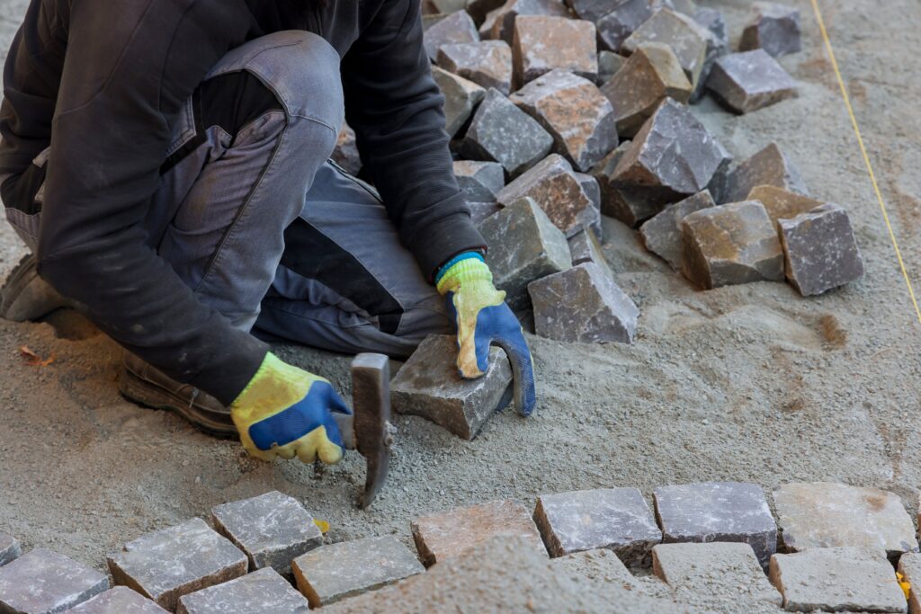 Road master in gloves lays paving stones in layers stone road by professional worker using stones and hammer - hardscaping services